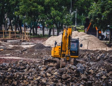 A yellow excavator working at an urban construction site surrounded by rocks and trees.