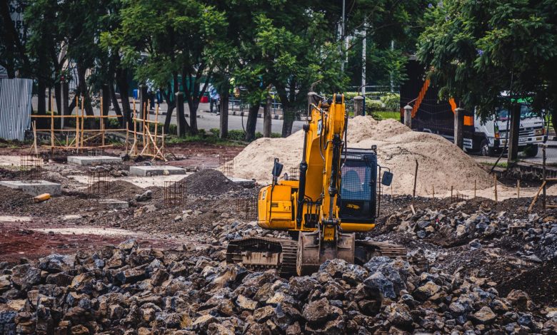A yellow excavator working at an urban construction site surrounded by rocks and trees.