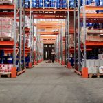 A spacious industrial warehouse with stocked shelves and a worker in protective gear.