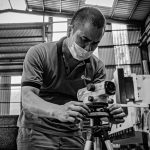 Black and white image of a man adjusting equipment in a warehouse setting, wearing a face mask.