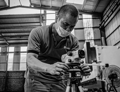 Black and white image of a man adjusting equipment in a warehouse setting, wearing a face mask.