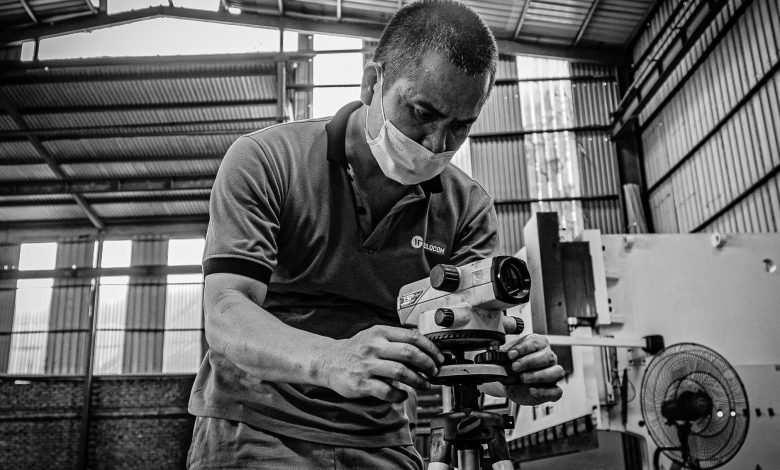 Black and white image of a man adjusting equipment in a warehouse setting, wearing a face mask.
