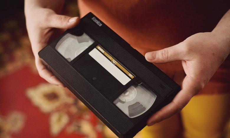 A person holds a vintage VHS tape against a warm, patterned background, evoking nostalgia.