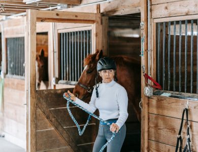 An adult woman holds a horse in a rustic wooden stable, emphasizing equestrian lifestyle.