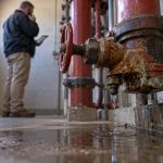 A regional property facilities manager stands in a cramped apartment-building sprinkler riser room, calling a shortlisted