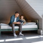 A father and son sharing a bonding moment while reading a book in an attic bedroom.