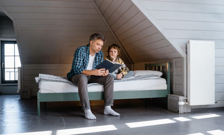 A father and son sharing a bonding moment while reading a book in an attic bedroom.