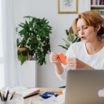 Woman reviewing bills at home desk with laptop and plants, managing personal finances.
