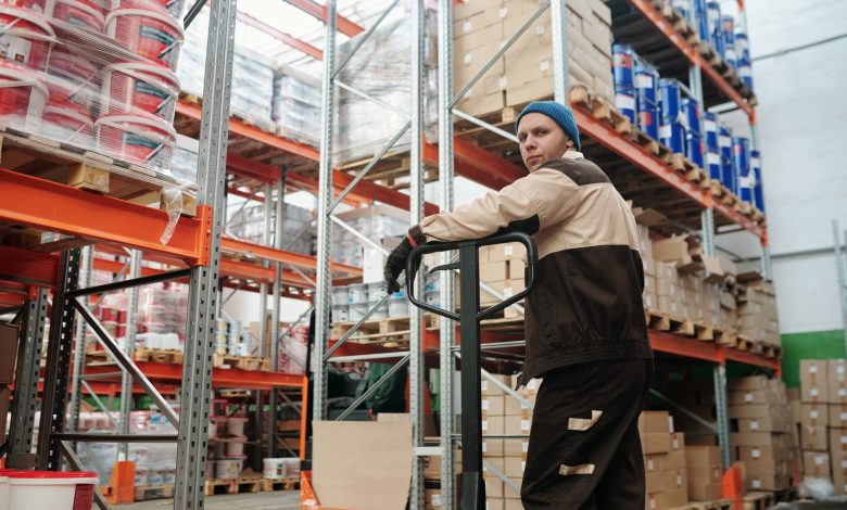 Warehouse worker operating a pallet jack among stacked boxes and metal shelves in an industrial setting.