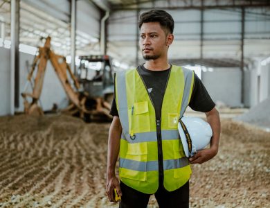 Portrait of a construction worker in safety gear holding a helmet inside an industrial setting.