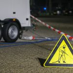 A construction warning sign and caution tape at night near a trailer, indicating ongoing work.