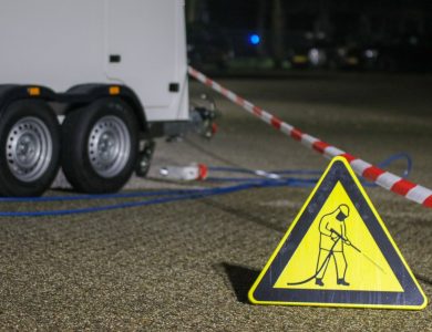A construction warning sign and caution tape at night near a trailer, indicating ongoing work.