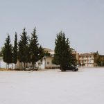 Wide shot of an empty urban lot with buildings and tall trees under a clear sky.