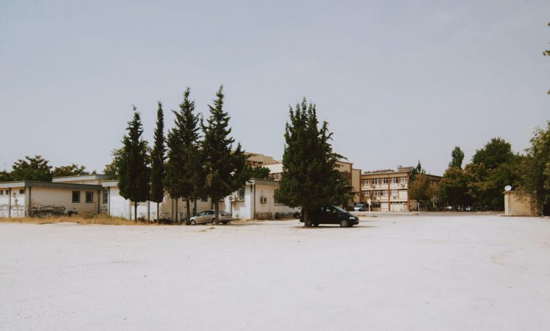 Wide shot of an empty urban lot with buildings and tall trees under a clear sky.