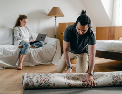 Fit ethnic male in wristwatch laying carpet on parquet while barefoot woman browsing internet on portable computer sitting on cozy sofa in bedroom