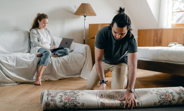 Fit ethnic male in wristwatch laying carpet on parquet while barefoot woman browsing internet on portable computer sitting on cozy sofa in bedroom