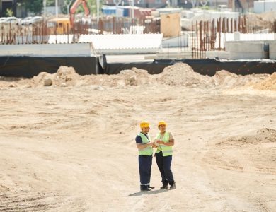 Two workers discussing plans on a sandy construction site, wearing safety gear.