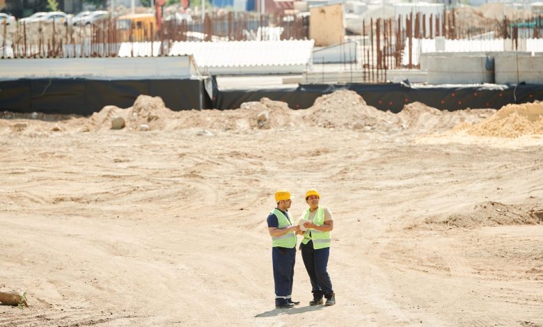 Two workers discussing plans on a sandy construction site, wearing safety gear.
