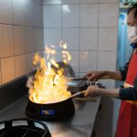 An Asian chef wearing a face mask cooks a flaming dish in a commercial kitchen.
