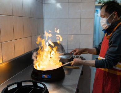 An Asian chef wearing a face mask cooks a flaming dish in a commercial kitchen.