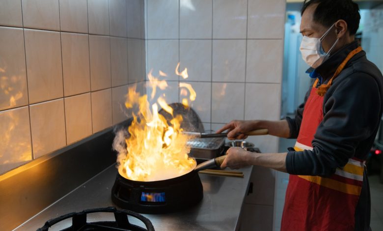 An Asian chef wearing a face mask cooks a flaming dish in a commercial kitchen.