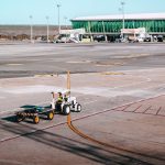 A luggage cart on the airport tarmac near a modern terminal building under clear skies.