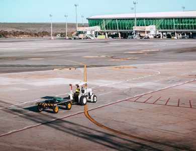 A luggage cart on the airport tarmac near a modern terminal building under clear skies.