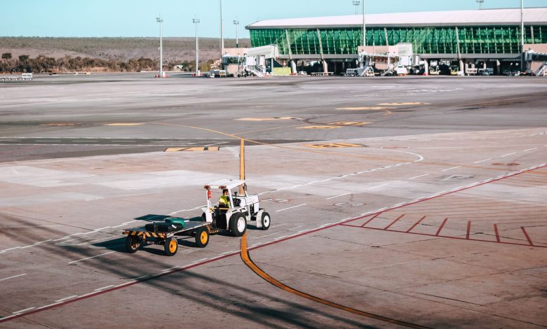 A luggage cart on the airport tarmac near a modern terminal building under clear skies.