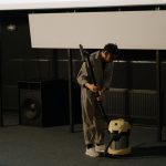 A person in uniform using a vacuum cleaner inside an auditorium with a visible speaker.