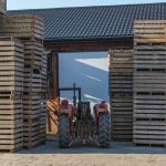 Red tractor positioned among towering stacks of wooden pallets outside a warehouse.